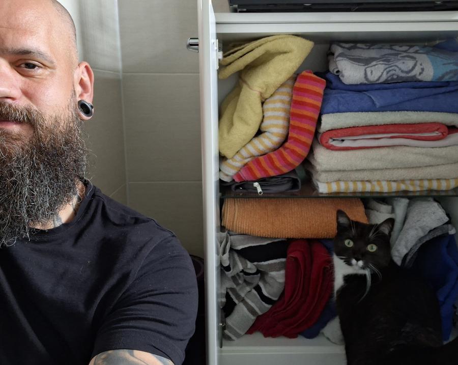 Ein bärtiger Mann sitzt vor einem geöffneten Schrank voller Handtücher. Kater Cooper sitzt daneben und findet die Falttechnik des Mannes eher grauenvoll.