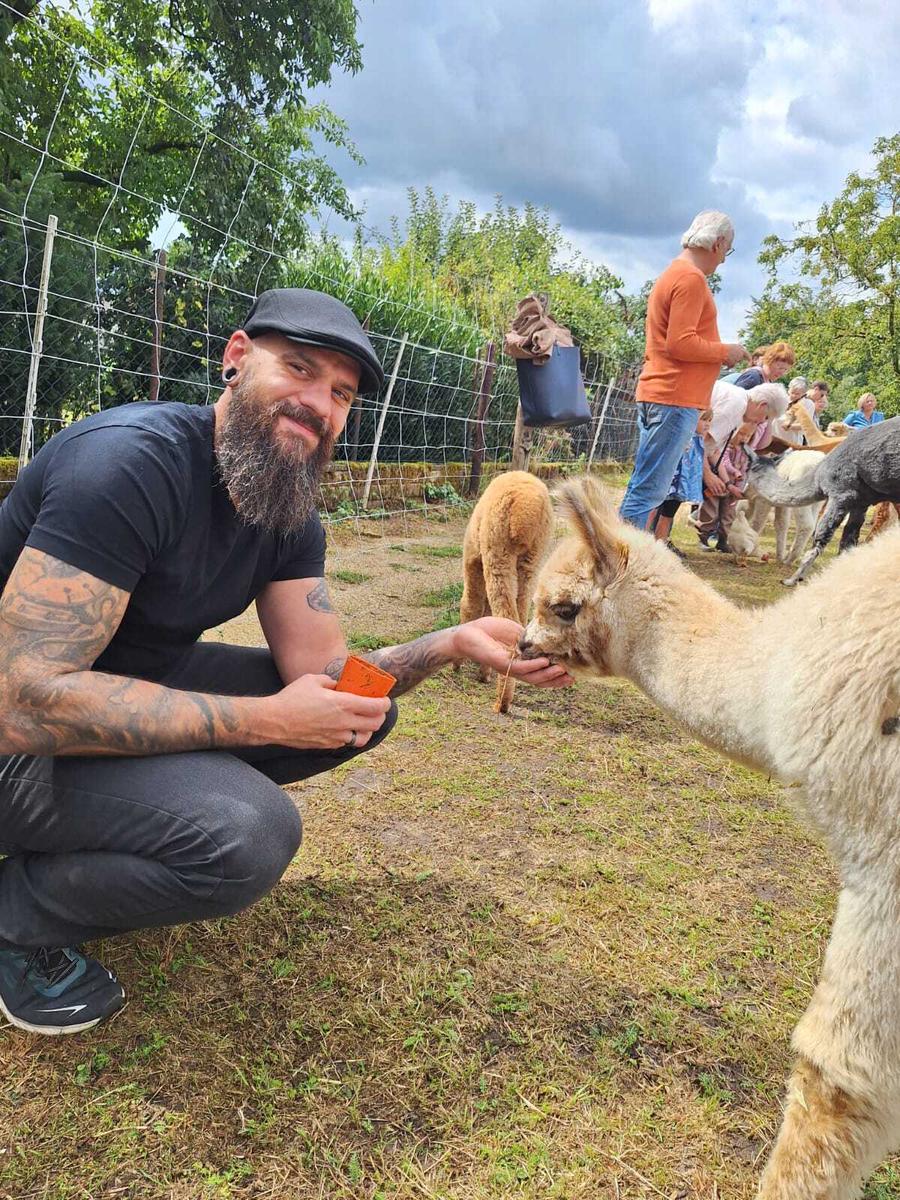 Ein Foto von einem Christian und dem Alpaka, das aus seiner Hand frisst. Das Alpaka hat eine modische Kurzhaarfrisur und scheint im Alter der Gen-Z anzugehören. Seine Hobbies sind Shisha-Bar und Pumpen im Fitty. Am liebsten hört es Oldies von Skrillex, wenn es mit dem Scooter über die Weide brettert.